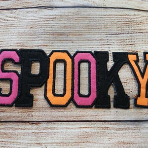 May include: A close-up of a felt patch that reads "SPOOKY" in black, orange, and pink letters. The letters are outlined in black and have a slightly raised texture.