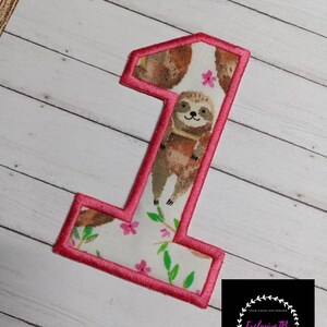 May include: A pink felt number one with a sloth and floral pattern. The number is surrounded by a pink border.