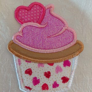 May include: Embroidered cupcake applique with pink glitter frosting and a pink polka dot heart. The cupcake liner is white with red and pink hearts and gold flecks. The applique is outlined in white.