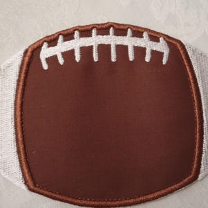 May include: Embroidered brown football-shaped patch with white stitching details. The patch features white stripes on the sides and white stitching across the center, resembling the laces of a football. The patch is likely for sports-themed crafts.
