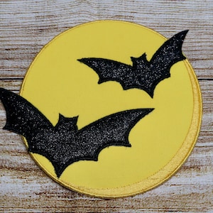May include: Two black bats are silhouetted against a yellow circle, resembling a full moon. The bats are facing different directions, with one bat flying above the other. The background is a light brown woodgrain surface.