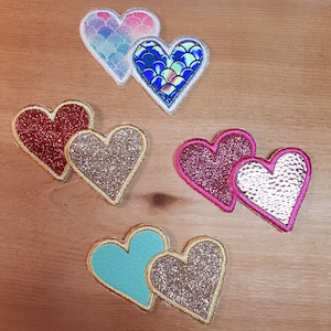 Puede incluir: Cinco pares de parches de fieltro en forma de corazón en varios colores y patrones. Los parches son de varios colores, incluyendo rojo, rosa, azul, turquesa y dorado. Algunos de los parches tienen brillo o un patrón de escamas.