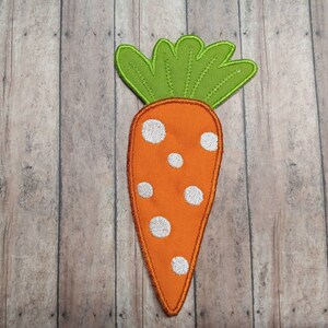 May include: A felt orange carrot with white polka dots and a green felt top. The carrot is on a wood background.