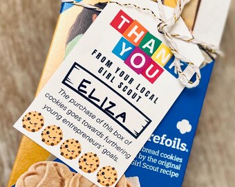 May include: A box of Girl Scout cookies with a tag that says "Thank You" and "From Your Local Girl Scout". The tag is personalized with the name "Eliza". The box contains 9 oz (255g) of cookies.