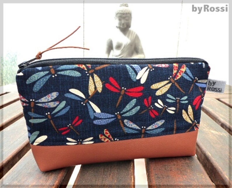 May include: A blue zippered pouch with a brown leather bottom. The pouch is decorated with a repeating pattern of colorful dragonflies. The text "by Rossi" is printed on the side of the pouch.