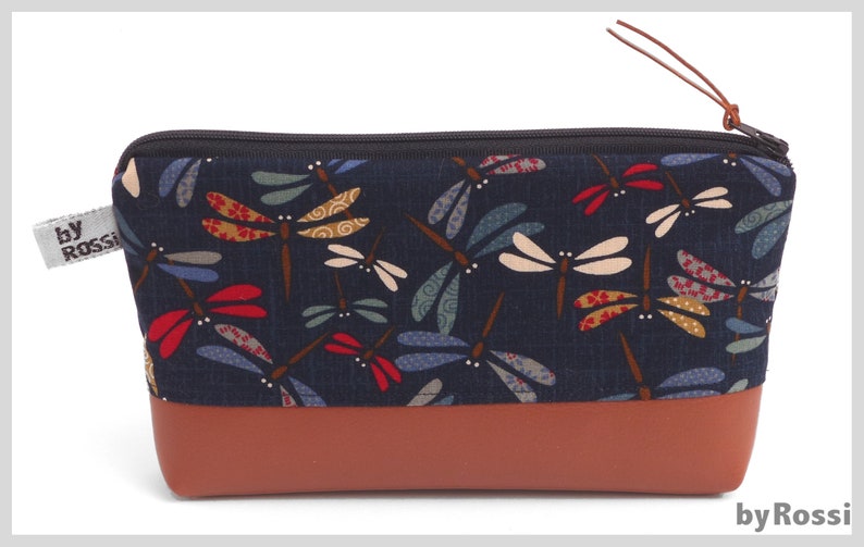 May include: A navy blue zippered pouch with a brown leather bottom. The pouch is decorated with a repeating pattern of colorful dragonflies.