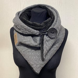 Könnte beinhalten: Grau-schwarzer Schal mit Knopfverschluss. Der Schal hat ein gestepptes Muster auf der Außenseite und ein weiches, schwarzes Futter. Ein großer, dunkler Knopf sichert den Schal, und ein kleines Etikett ist sichtbar.