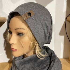 Könnte beinhalten: Graue Mütze mit einem braunen Lederpatch, auf dem "Good Day" steht, und einem schwarzen Knopf. Die Mütze wird mit einem grauen Schal getragen.