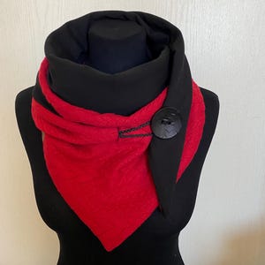 May include: A black and red scarf with a button fastening. The scarf has a textured red section and a black hood. The button is large and dark, and the scarf is displayed on a black mannequin. The scarf is made of a soft material.