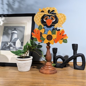 May include: A wooden crow figurine wearing a yellow plaid hat and overalls, holding sunflowers. The crow is mounted on a wooden candlestick. A framed black and white lighthouse print and a small potted plant are also visible.