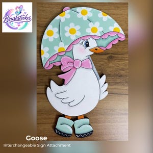 May include: A flat wooden cutout of a white goose wearing light green rain boots and a pink bow. The goose holds a light green umbrella decorated with a white daisy pattern and a pink scalloped edge. The goose has an orange beak and blue eyes.