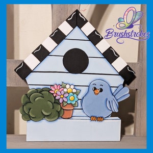 May include: A light blue wooden birdhouse with a black and white chequered roof. A blue bird, green bush, and potted flowers are attached to the front. The birdhouse has a round black opening. The text "Brushstrokes" is in the upper right corner.