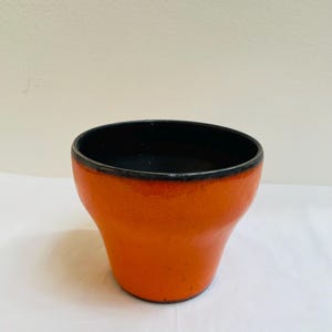 Op de afbeelding: Een kleine, oranje keramische pot met een zwarte rand. De pot heeft een getextureerd oppervlak.