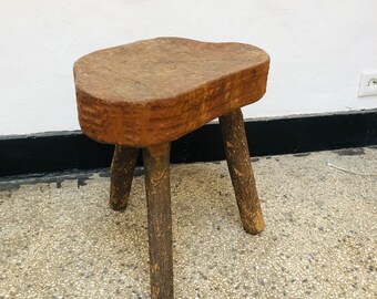 Puede incluir: Un taburete de madera rústico con una parte superior ancha y desigual y tres patas robustas. El taburete está hecho de madera natural con un acabado marrón.