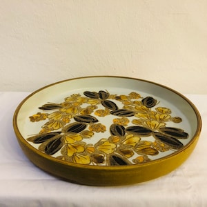 Può includere: Un grande vassoio rotondo in ceramica con un bordo marrone e un centro bianco. Il vassoio è decorato con un motivo floreale in tonalità di giallo, marrone e nero.