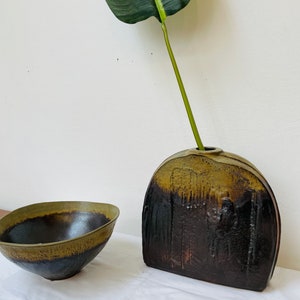 May include: A dark green and brown ceramic vase with a rounded, arched shape. A single green leaf is in the vase. A smaller, dark green and brown ceramic bowl is also in the image.