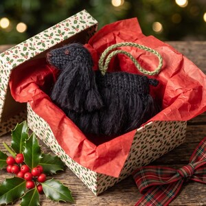 May include: A black crocheted dog ornament with long, textured yarn ears and a green braided hanging loop, nestled inside a gift box lined with red tissue paper, accompanied by a sprig of holly and a red and green plaid ribbon.