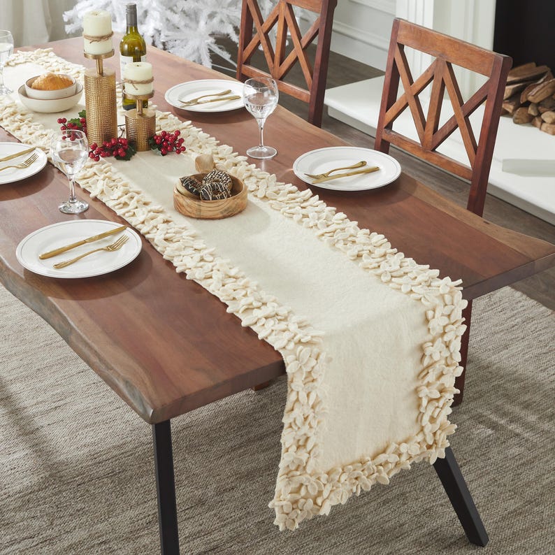 May include: A white table runner with a fluffy, textured border. The runner is laid on a wooden table with a natural edge. The table is set with two place settings, each with a white plate, gold silverware, and a wine glass.