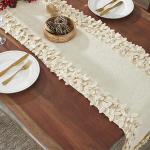 May include: A white table runner with a decorative border of white felt flower shapes. The runner is on a wooden table with a plate and silverware.