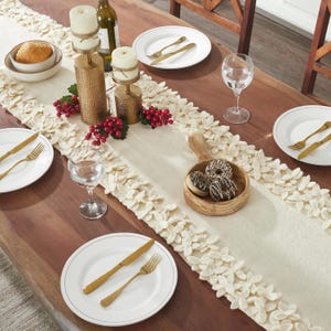 May include: A white table runner with a fluffy, leaf-shaped trim. The runner is decorated with red berries and gold candle holders. There are two white plates with gold cutlery, a glass of water, and a wooden bowl with donuts on the table.