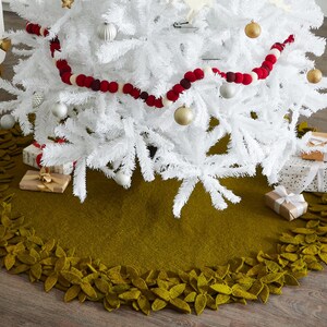 May include: A green felt Christmas tree skirt with a leafy border. The skirt is decorated with felt leaves and is placed under a white artificial Christmas tree.