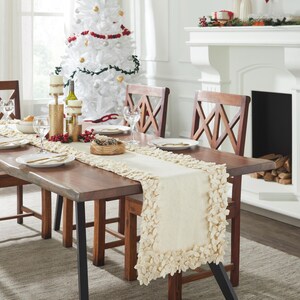 May include: A white table runner with a ruffled edge, placed on a wooden table with a Christmas tree in the background.