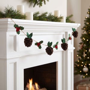 May include: A festive garland featuring alternating felt brown pinecones and green holly leaves with small red berries, all strung together on a thin twine cord. The decorative piece is draped across the front of a white fireplace mantel.