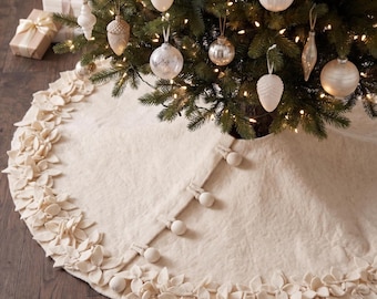 May include: A cream-colored felt Christmas tree skirt featuring a border of layered fabric leaves and a front closure secured with four round fabric-covered buttons. The skirt is positioned beneath a decorated evergreen tree.