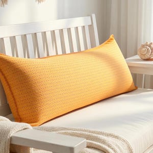 May include: A long, rectangular yellow pillow with a stitched pattern rests on a white bench. The pillow is a warm, golden color and appears soft. A white blanket is draped over the bench's arm. A seashell sits on a small side table.