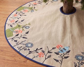 May include: A circular beige tree skirt featuring embroidered green deer, blue birds, and orange floral motifs. The fabric edge is finished with a dark blue trim, and a central circular opening is reinforced with matching blue piping.