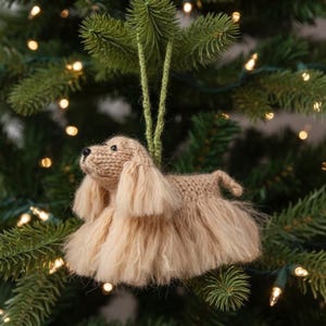 May include: A tan knitted dog ornament featuring long, fluffy ears and a textured body. The dog has a small black nose and dark eyes, and it is suspended by a green yarn loop for hanging. The figure is crafted with a soft, fuzzy yarn texture.
