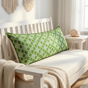 May include: A rectangular decorative pillow with a green and white pattern, featuring a green border. The pillow is placed on a white bench, with a cream-colored blanket draped over the arm. A seashell sits on a small side table.