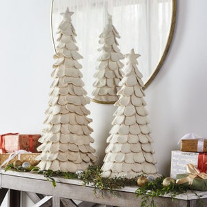 May include: Three white felt Christmas trees with a star on top, decorated with greenery and gold ornaments. The trees are on a wooden table.