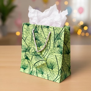 May include: A green gift bag with a botanical pattern of dark green pine needles and cones. The bag has a light pink rope handle and is filled with white tissue paper. The bag is sitting on a wooden surface.