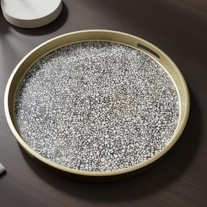 May include: A round decorative tray with a gold-colored rim and handles. The tray's surface features a black and white terrazzo pattern. The tray is designed for serving or display purposes.
