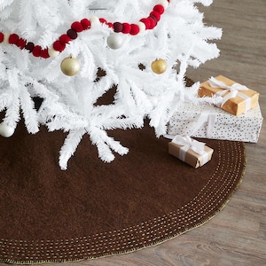 May include: A brown felt Christmas tree skirt with a gold stitched border. The skirt is decorated with a white Christmas tree and red and white felt ball garland.