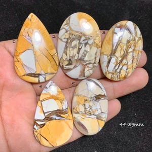 Può includere: Cinque cabochon di pietre preziose lucide nei toni del giallo, marrone e bianco. Le pietre variano nella forma, tra cui ovale e a goccia. Il testo "44-39mm" è visibile. Queste pietre sono probabilmente per la creazione di gioielli o la collezione.