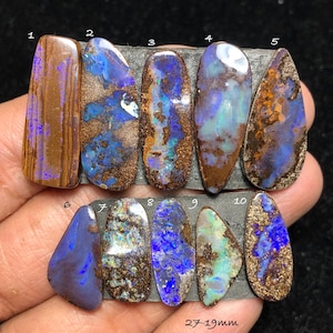 Puede incluir: Una colección de diez piedras preciosas de ópalo pulidas, cada una con patrones y colores únicos. Los ópalos varían en forma y tamaño, con una mezcla de tonos marrones, azules y verdes. El texto "27-19mm" es visible, indicando el tamaño de las piedras.