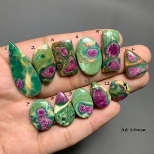 Puede incluir: Once cabujones pulidos de fuchsite verde con cristales de rubí magenta incrustados, numerados del 1 al 11. Las piedras presentan formas variadas que incluyen gota, óvalo y rectángulo, con tamaños que van de 36mm a 14mm según se indica.