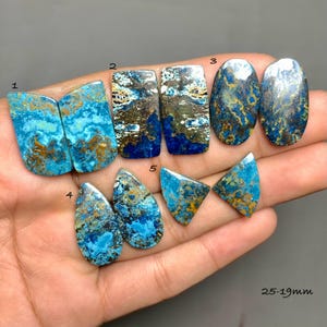 Puede incluir: Una colección de piedras de azurita pulidas en varias formas, incluyendo rectangulares, ovaladas y en forma de lágrima. Las piedras muestran una vibrante mezcla de tonos azules, marrones y dorados. La imagen incluye el rango de tamaño de las piedras, que es de 25-19mm.