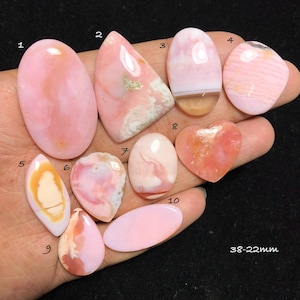 May include: Ten polished pink opal cabochons in various shapes including oval, heart, and teardrop, numbered 1 through 10. The stones display shades of light pink, peach, and white with natural banding and inclusions. Text 38-22mm is visible.