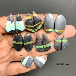 Op de afbeelding: Een collectie gepolijste cabochon edelstenen met een zwarte en grijze matrix en levendige turkooiskleurige aders. De stenen zijn genummerd van 1 tot 7 en bevatten diverse vormen zoals druppels, ovalen en harten, met tekst voor 23-15mm.