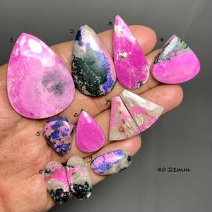 Puede incluir: Una colección de nueve piedras preciosas pulidas en varias formas, incluyendo gota, ovalada y triangular. Las piedras presentan patrones moteados en rosa vibrante, azul profundo, negro y blanco. Cada piedra está numerada del 1 al 9, con el texto 40-21mm.
