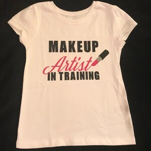 Pode incluir: Camiseta branca com texto preto que diz "MAKEUP Artist IN TRAINING" e um gráfico de batom rosa.