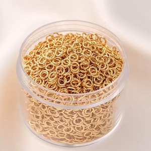 Könnte beinhalten: Ein durchsichtiger Kunststoffbehälter gefüllt mit zahlreichen kleinen, goldfarbenen Sprungringen. Die Ringe sind dicht gepackt, reflektieren das Licht und erzeugen einen schimmernden Effekt. Ideal für die Schmuckherstellung und Bastelarbeiten.