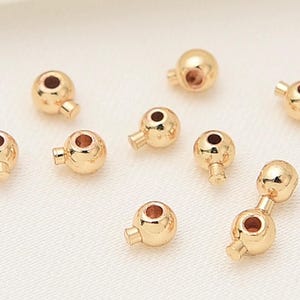 May include: Several small, gold-coloured metal beads are displayed. Each bead is spherical with a hole and a small cylindrical extension. The beads are scattered on a light-coloured surface, possibly for crafting or jewellery making.
