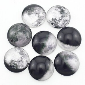 Puede incluir: Ocho cabujones redondos de vidrio con una imagen de la luna impresa en ellos. Los cabujones son negros y blancos con un tinte gris y verde.