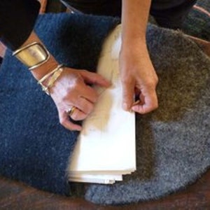 May include: A person is sewing a piece of fabric to a stack of papers with a needle and thread. The fabric is a dark blue color and the papers are white. The person is wearing a silver bracelet on their wrist.