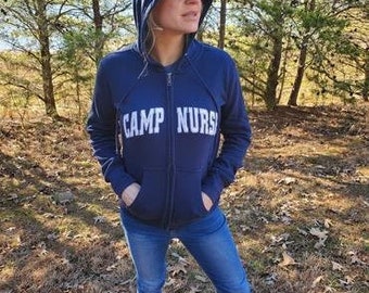 May include: A navy blue zip-up hoodie with a white hood and white text that reads "CAMP NURS" on the front. The person wearing the hoodie is standing in a wooded area.