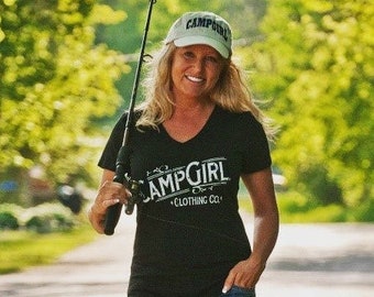 May include: Black V-neck t-shirt with the text "CAMPGIRL CLOTHING CO." and a light green cap with "CAMPGIRL" printed on it. The person is holding a fishing rod and wearing blue jeans. The background is a blurred outdoor scene.